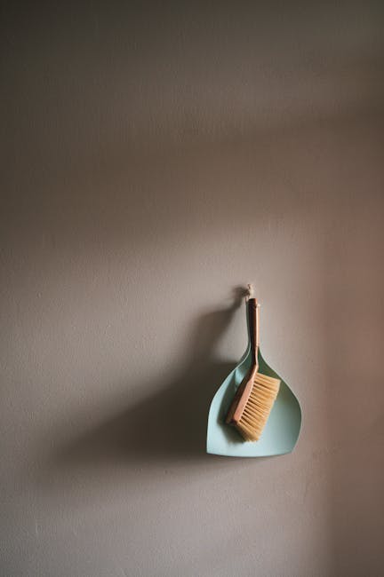 A beige wall with a small, light blue wall hook holding a wooden cleaning brush with light brown bristles and a light blue dustpan. The dustpan and brush are positioned vertically on the wall, with th